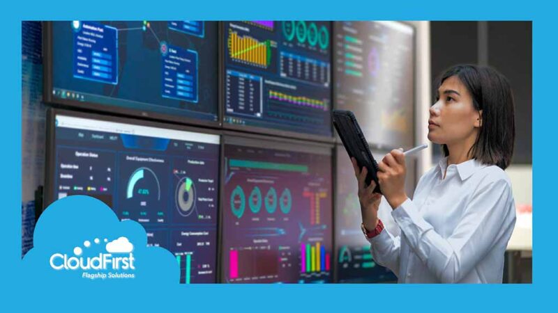 A woman writing on a tablet and standing in front of a series of data dashboards